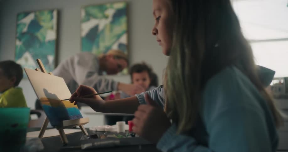 A girl sits in a kitchen at home painting vibrant landscapes on small canvases as sunlight streams through the windows
