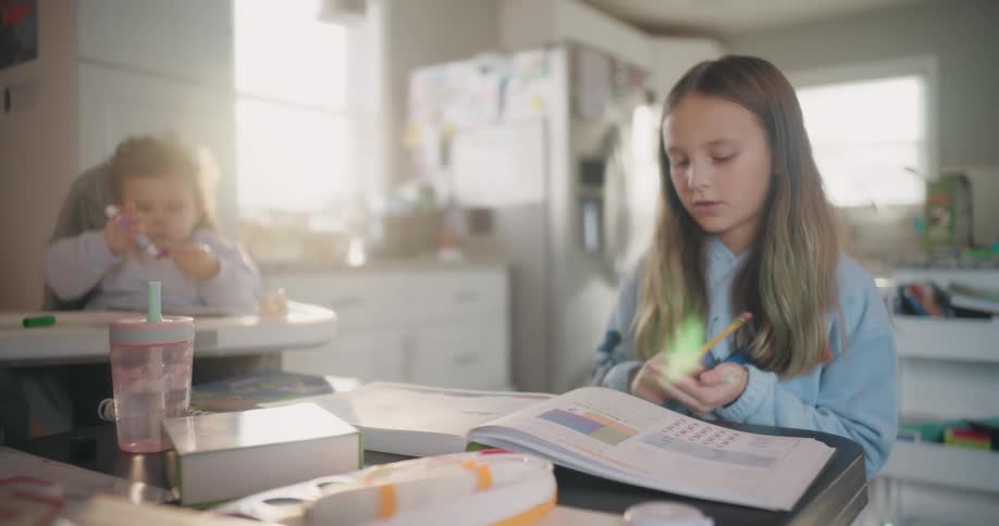 A girl doing math schoolwork in a kitchen next to a toddler in a high chair