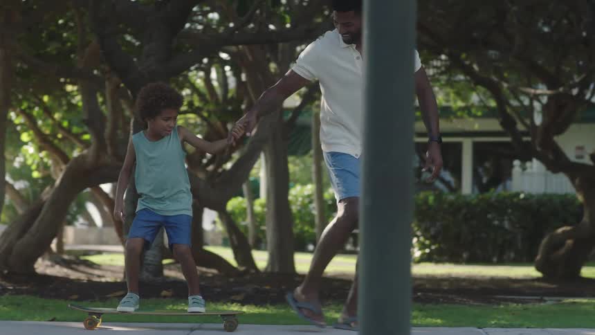 A father teaches his son to ride a skateboard in the park on a sunny day