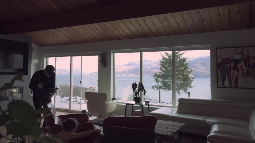 A man uses a camera to film a girl playing with chairs in the living room of a lake house