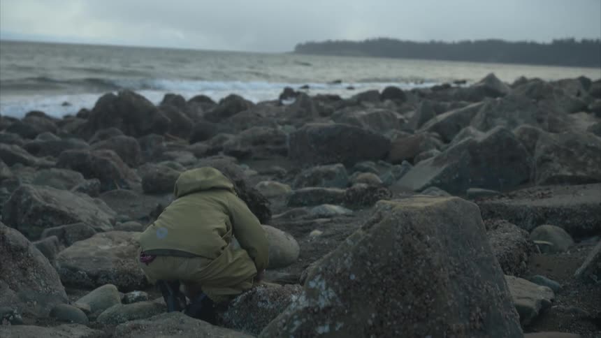 A child navigates a rocky shoreline in a beige jacket and boots leaning down to pick up a stone among scattered rocks while gentle waves roll in beneath a muted overcast sky