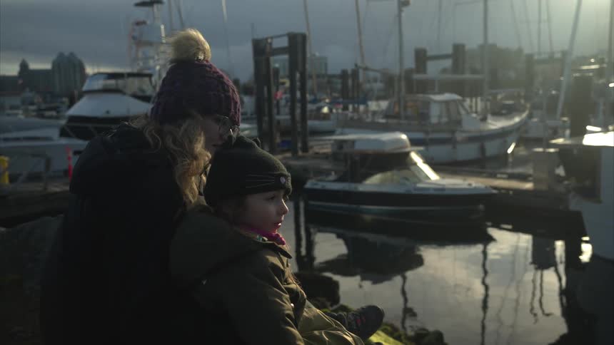 A mother and her daughter sit looking at boats on a marina on a cold overcast day