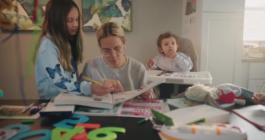 Mother sitting at a table paying attention to a girl with an open book and around her a baby in the background and another boy writing in a notebook