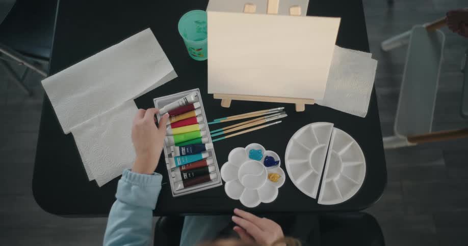 A person puts tubes of paint on a tray of a table with canvas and an easel