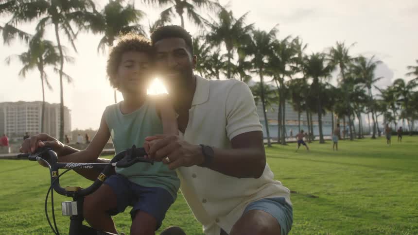 A father is teaching his song how to ride a bike in a popular park encouraging him and giving him a tender kiss on the cheek as people play frisbee in the background and palm trees sway gently in the wind in front of the midmorning sun