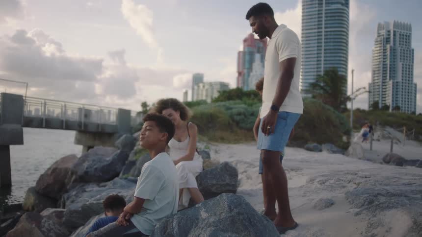 A family is gathered on a rocky shoreline with a waterfront city in the background at dusk