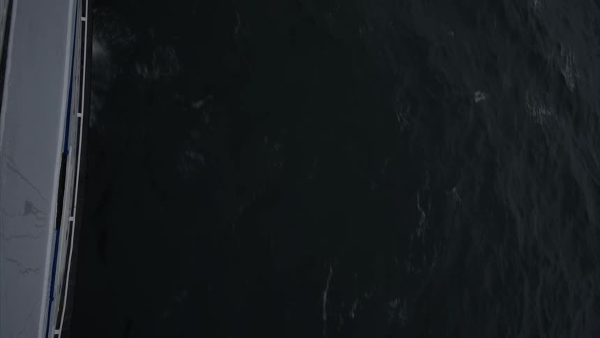 Viewed from above the side of a ship slices through dark ocean water leaving a trail of foamy white waves that churn and crash along its hull starkly contrasting against the deep surface below