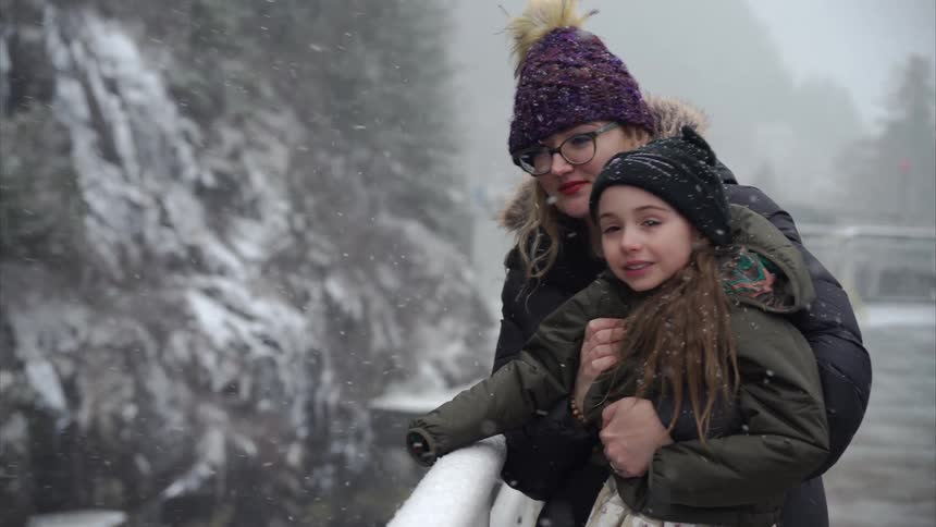 A mother holding her daughter in her arms both dressed in winter clothing stand on the snowy deck of a ship gazing overboard