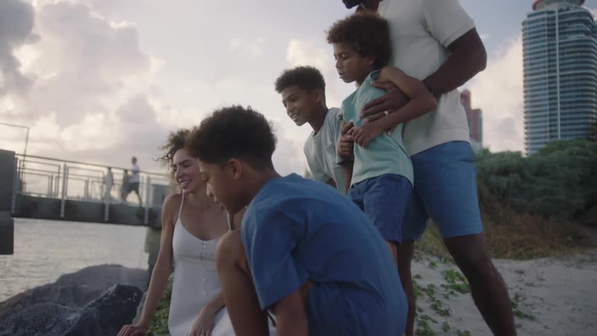 A family is on the rocks near sand with a bridge and building in the background