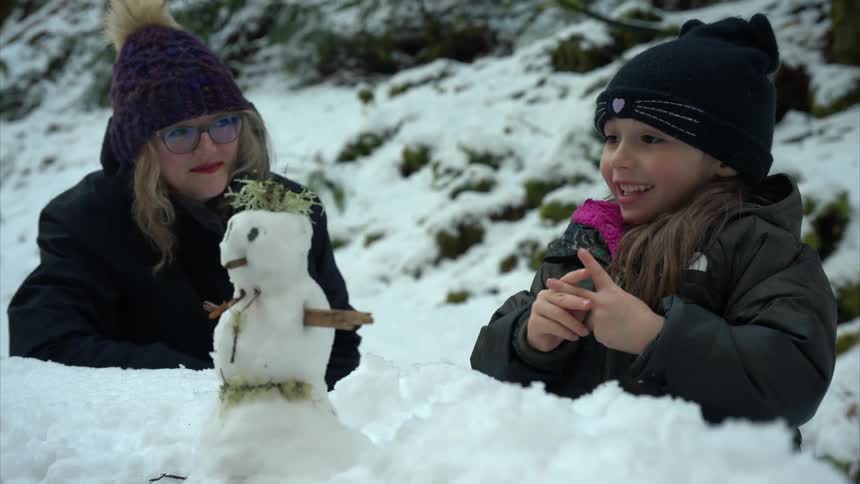 A mother and daughter in winter clothes build a small snowman in a snowy landscape