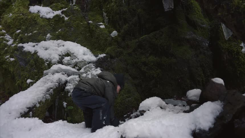 The girl is climbing up the small bank at the base of the cliff and she picks up a chunk of ice and turns and smiles at someone who is not visible