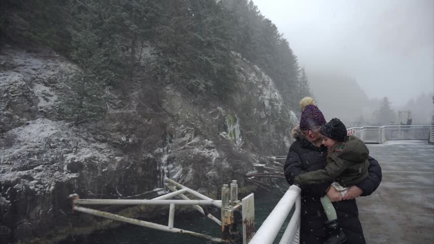 Mother holding her daughter on her lap while standing outside on a moving ship on a snowy day