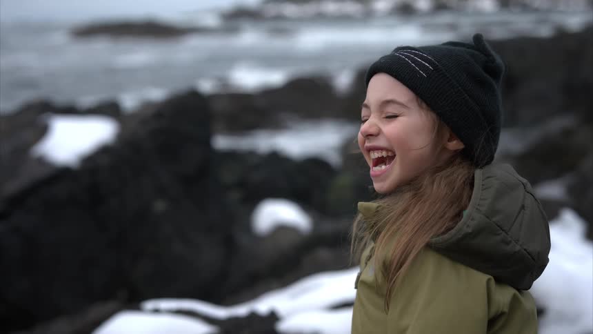 A little girl smiles as she stands near water on snowy rocks