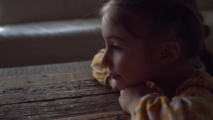 The little girl is resting her chin on her hands that are resting on the table and she is staring ahead
