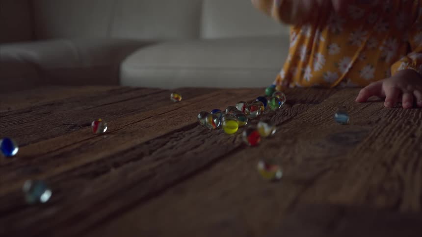 A child in a yellow floralpatterned outfit playing with colorful glass marbles on a rustic wooden table with a beige couch in the background