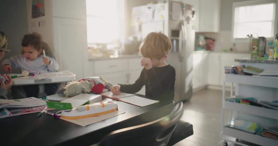 A boy looks upset doing his homework as he sits at a table with his mom and brother