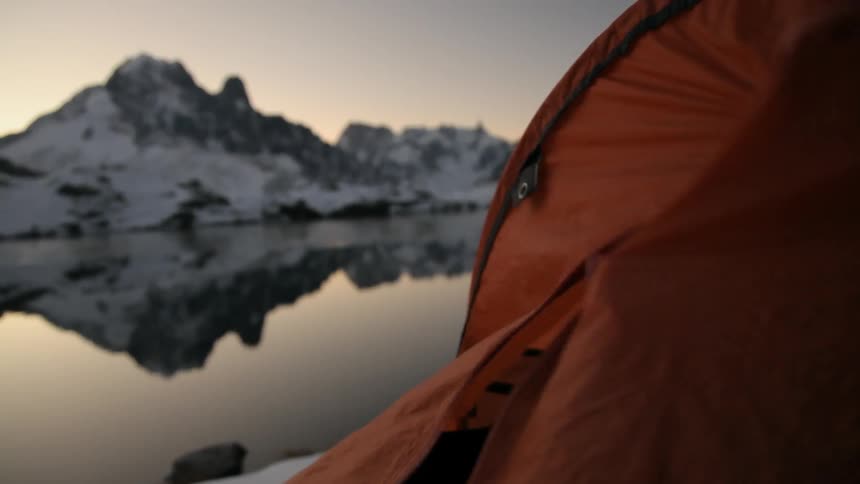 Person unzipping a tent near a lake and snow covered mountains