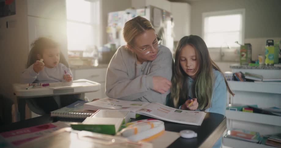 A mother helps her daughter with homework in the kitchen while a baby sits nearby on a high chair eating