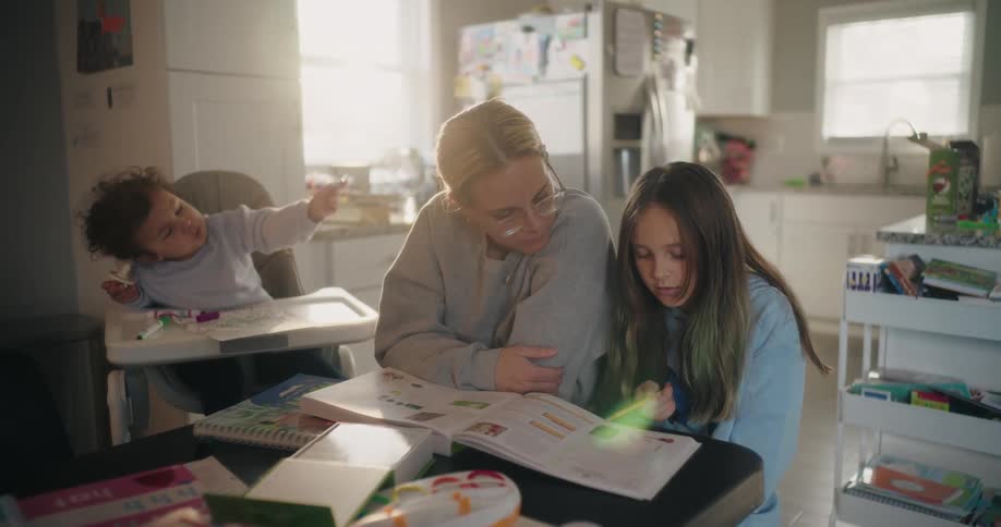 A mother helps her daughter with homework while a baby sits in a highchair
