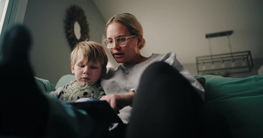 The mother and the little boy are sitting on the couch and she is reading to him and showing him something in a book