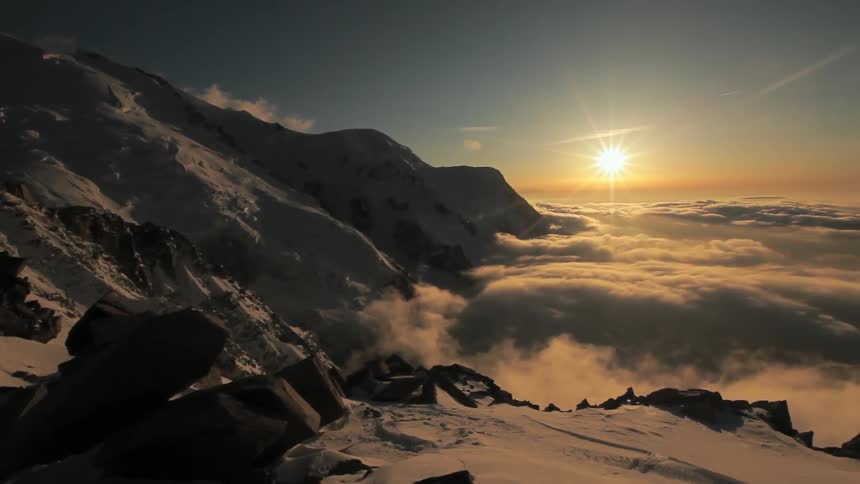 The sun shines above clouds that hang near the peaks of a mountain range
