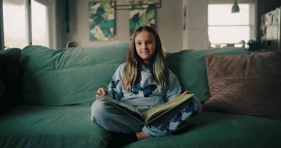 A girl with long hair sits on a green couch in a living room a book open on her crossed legs