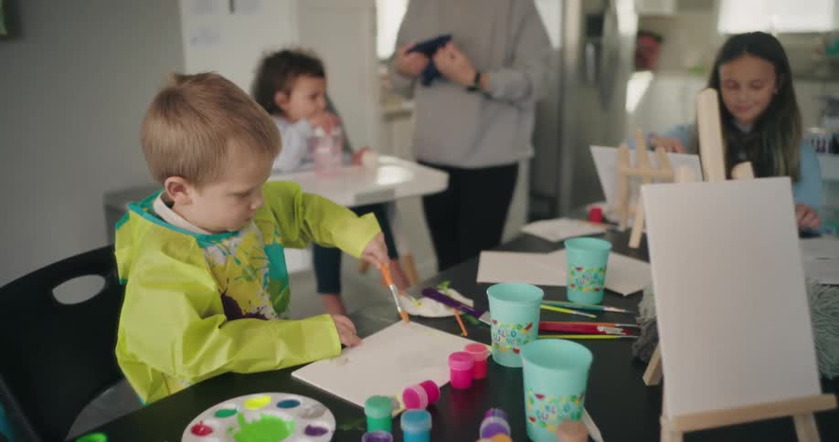 A group of children and an adult are engaged in a painting activity at a table surrounded by colorful paint pots brushes and palettes