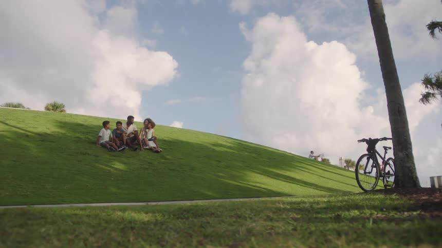 A family sits on a grassy hill under a bright sky with fluffy clouds while a bicycle leans against a tree trunk in the foreground