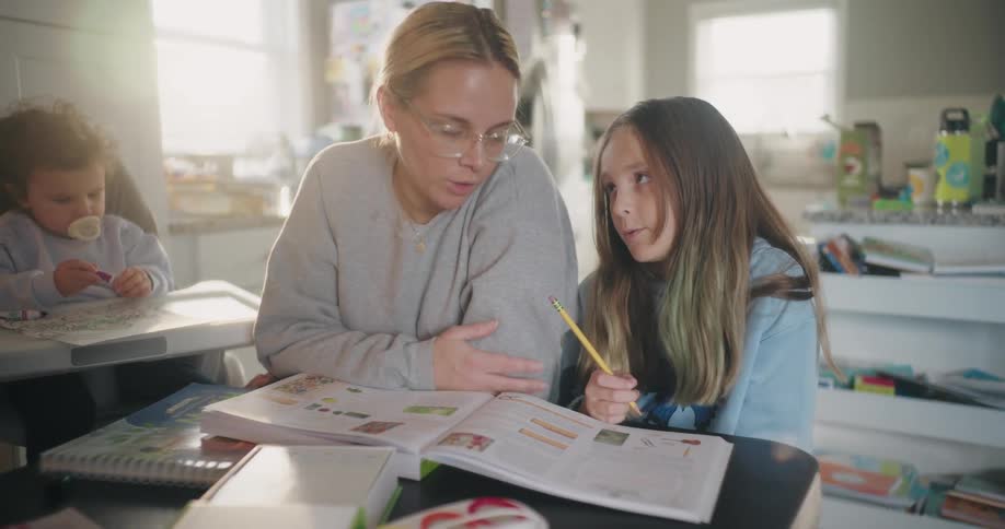 Mother teaching her daughter with an open notebook and the background of a baby and a child playing
