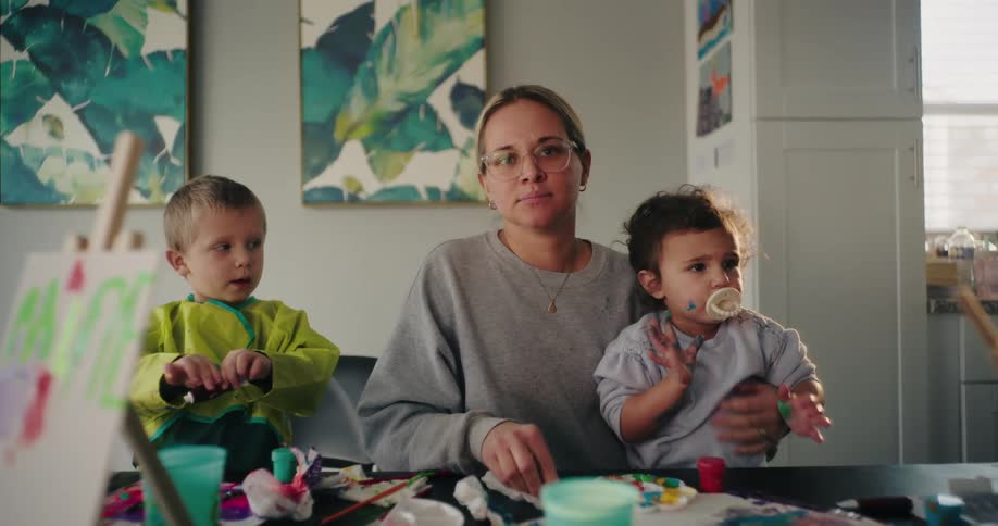 A woman and two young children are engaged in painting at a table covered with art supplies creating a colorful and playful scene