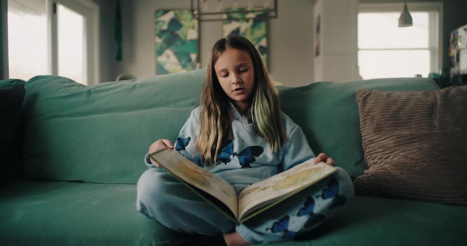 Girl sitting on the couch in the living room reading a book and a younger boy walking in front of her