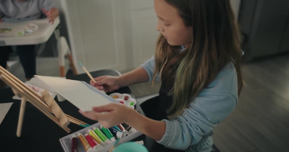 A girl paints on a canvas using a brush and a palette while her brother sits on a high chair in the background