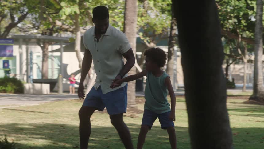 A father teaches his son to ride a skateboard on a sunny day in the park
