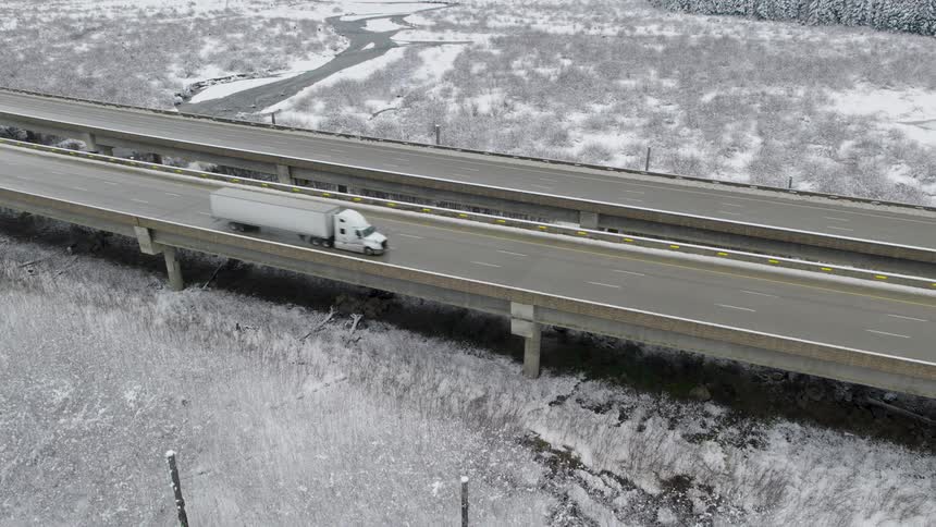 A white semi truck drives on a highway viaduct that spans a frosty winter landscape
