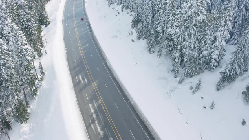 A red car climbs a freshly plowed highway surrounded by deep snow and frosted trees in crisp morning light