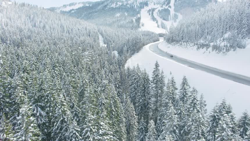 A black SUV travels up a winding road that cuts through a snowcovered valley flanked by snow laden evergreen trees