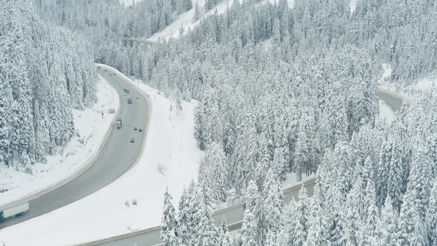 Big rigs and passenger vehicles make their way along a divided mountain pass highway surrounded by snow covered forest