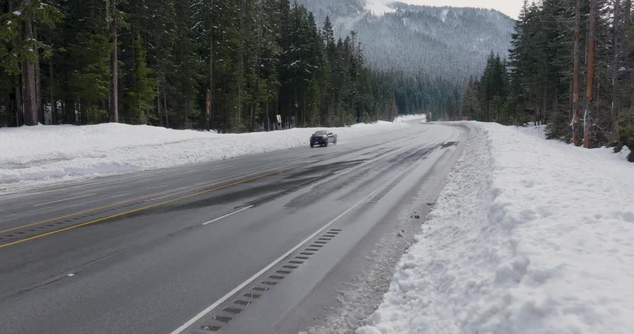 A black pickup truck travels on wet pavement along a highway through a snowcovered forest landscape