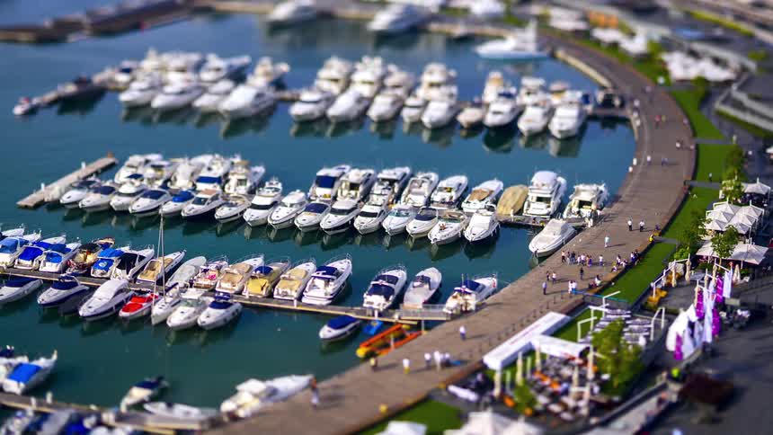 A tiltshift aerial view shows numerous yachts docked in a marina with a curving walkway