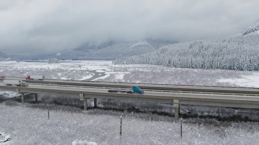 Cars and big rigs travel on a viaduct over a winter landscape with snow covered mountains in the distance