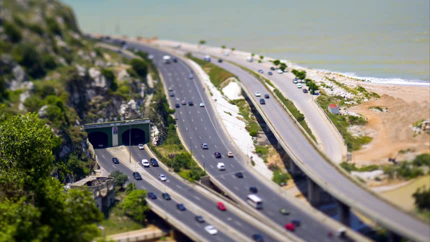 From an aerial vantage vehicles speed along a multilane highway near a hillside and coastline with a tiltshift effect lending a miniaturelike quality to the bustling traffic below
