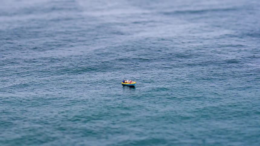 From an aerial vantage using a tiltshift filter a small boat traverses gently rolling waves in rapid timelapse its bright form highlighted against the vast expanse of blue water stretching out toward the horizon