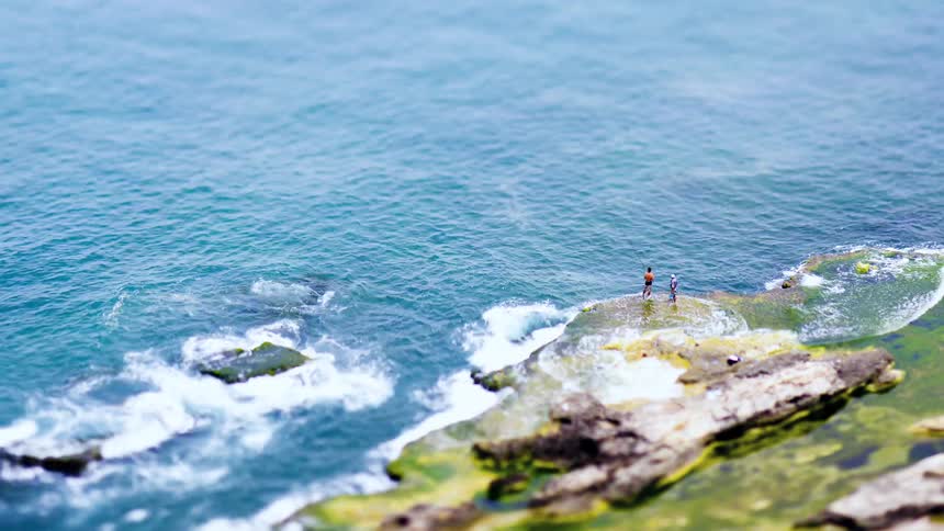 Two fishermen on top of a rock fishing on a tidal shore with some waves reaching their feet