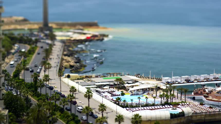 A coastal road lined with palm trees and cars runs alongside a waterfront area featuring a swimming pool sun loungers and a rocky shoreline with a distant pier