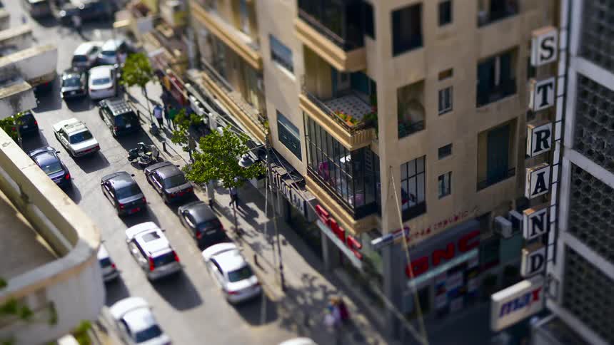 A tiltshift aerial view shows a city street with parked cars and buildings