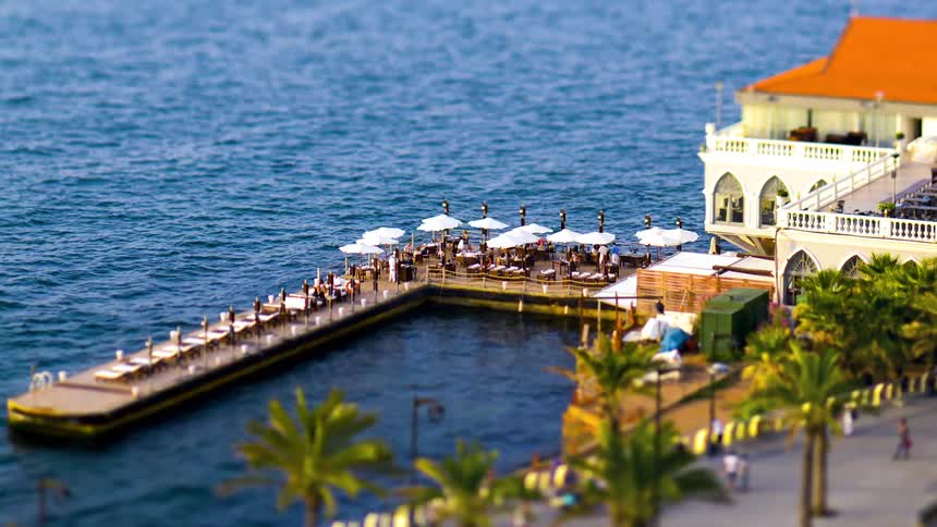 There are people walking everywhere along a pier that has umbrellas and a building at the beginning of it