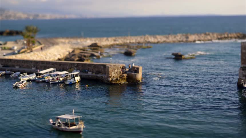 A tiltshift aerial view shows small boats docked in a harbor with a stone breakwater extending into the blue sea