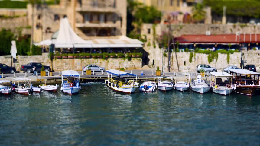 A line of small boats is moored along a pier with buildings in the blurred background