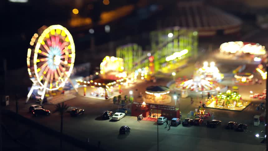 Under a nighttime sky a popup carnival with a ferris wheel and other attractions is shown in a tiltshift effect as people come and go in timelapse beneath dazzling lights and bustling activity