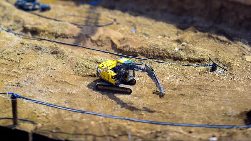 A yellow excavator digs in a dirt pit with black pipes running along the ground